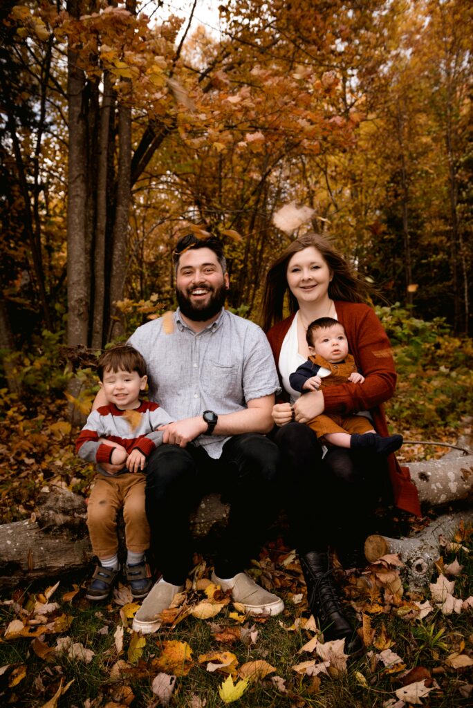 Smiling family sitting on a log in a forest, surrounded by autumn leaves.